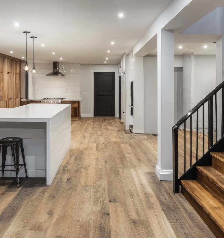 Modern basement renovation featuring an open-concept layout with a sleek kitchen island, wood-look flooring, recessed lighting, and a staircase with black metal railings. The design emphasizes functionality and contemporary style.