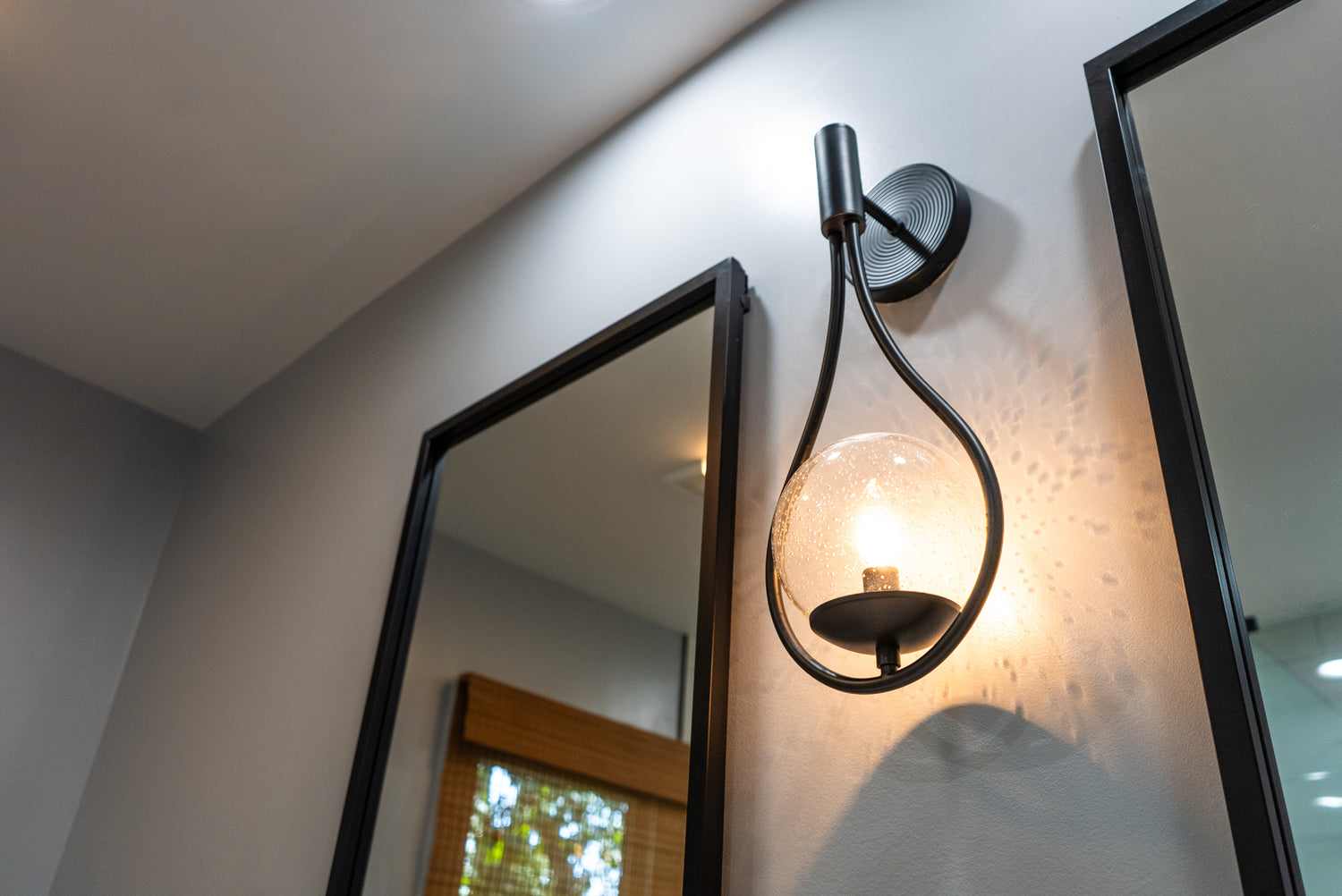 Black wall sconce with a curved teardrop arm and seeded-glass globe shade, mounted between two black-framed mirrors on a light gray bathroom wall