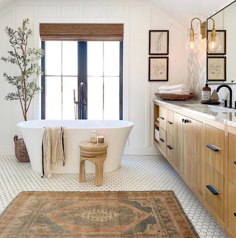 Bright bathroom with a freestanding white soaking tub, light wood vanity, black-framed window, hexagon tile floor, and a vintage patterned rug