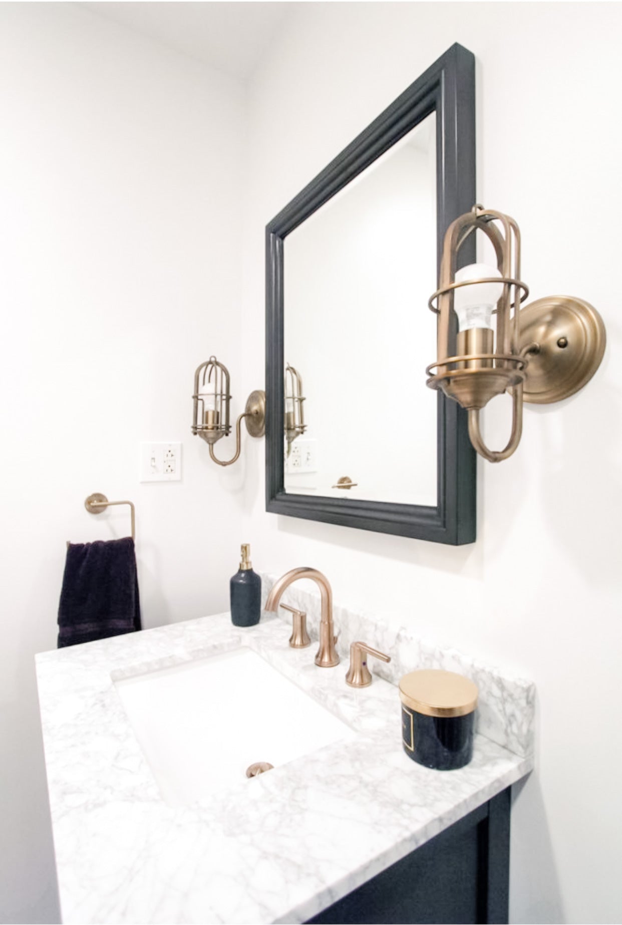 Navy vanity with marble countertop, brushed gold faucet and cage-style wall sconces flanking a dark-framed mirror in a bathroom