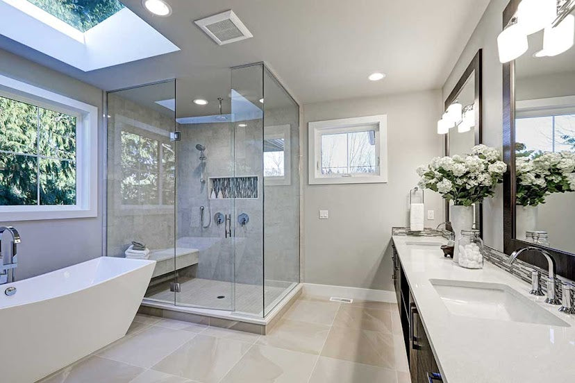 Bright, modern bathroom with a freestanding white bathtub beside a large window, a glass-enclosed walk-in shower with a built-in bench, and a double-sink vanity with white countertops and dark cabinetry. Neutral grey walls, large light-colored floor tiles, decorative flowers, and a skylight create an airy, spa-like atmosphere.