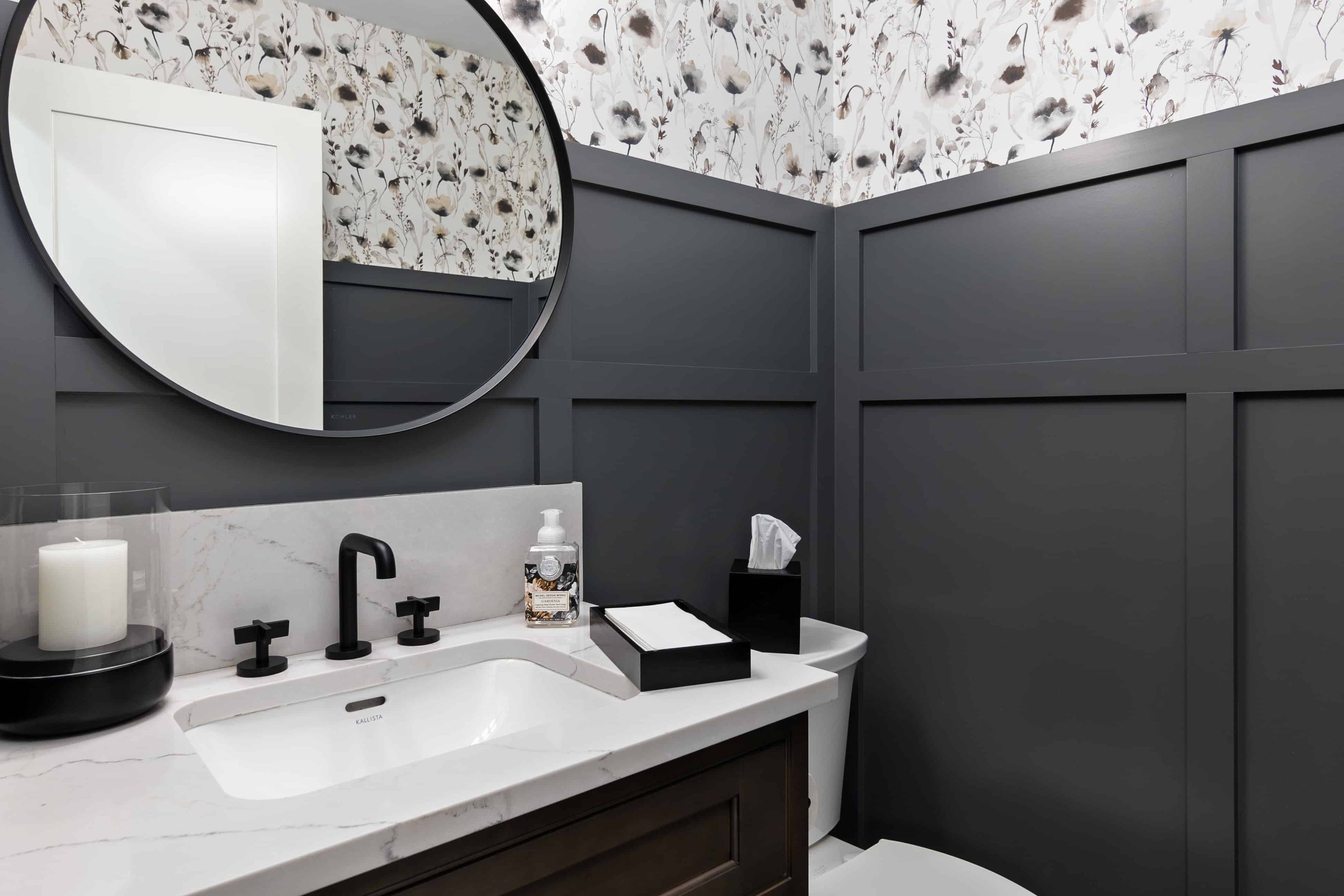 Modern powder room with grey board-and-batten wall paneling, white floral wallpaper above, and a white quartz vanity with subtle gray veining. A large round black-framed mirror hangs above the sink, which features matte black faucet fixtures. Decorative accessories include a candle, hand soap dispenser, tissue box, and folded guest towels. A white toilet is partially visible in the corner, creating a clean, elegant, and contemporary bathroom design.