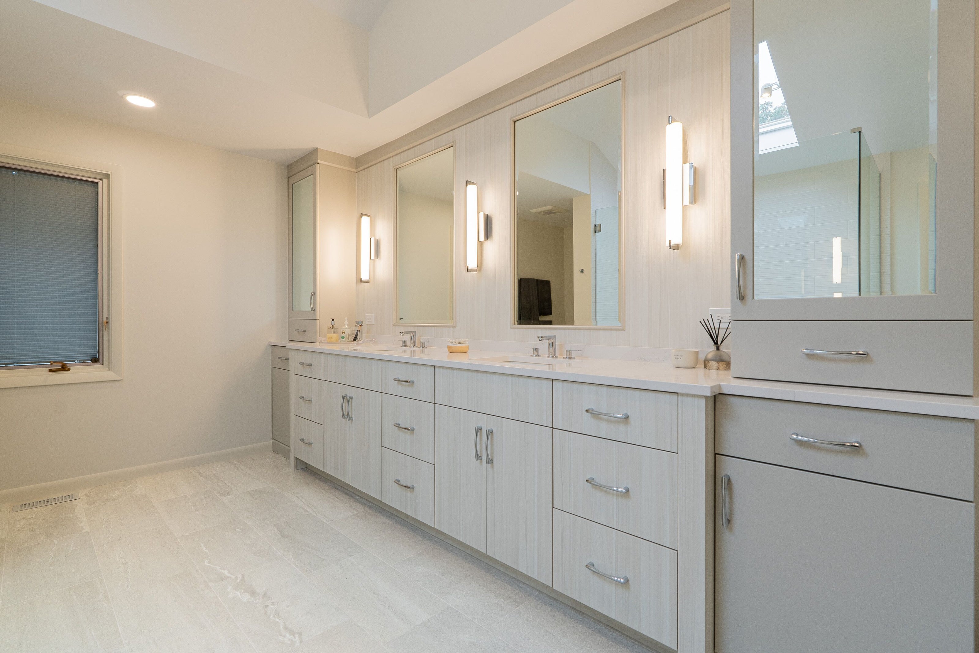 Spacious primary bathroom with double vanity in light wood-grain cabinetry, quartz countertops, and three framed mirrors under chrome sconces