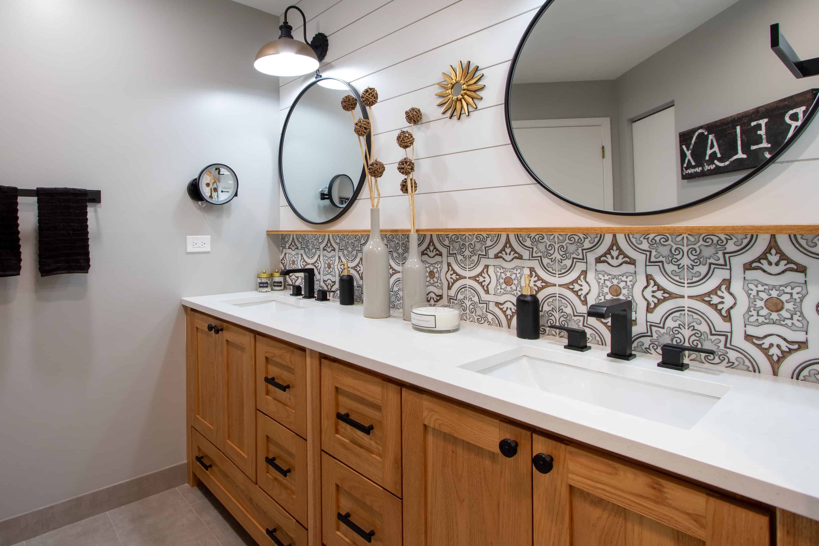 Contemporary double-sink bathroom featuring a long white countertop on a natural wood vanity with black hardware. Two large round black-framed mirrors hang above the sinks, complemented by black wall sconces and matte black faucets. A decorative patterned tile backsplash runs along the wall beneath white horizontal shiplap paneling. Neutral grey walls and flooring create a clean, modern look, while decorative vases, candles, and a gold sunburst wall accent add warmth and visual interest.